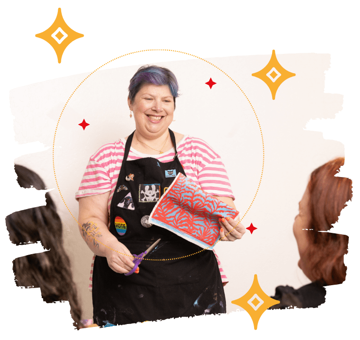 Art instructor Maria Pascoal wearing a black apron and holding a red and blue patterned paper during a workshop.
